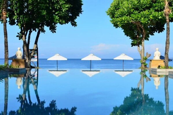 A clear pool with a reflection of trees and umbrellas, a statue, and a view of the sea.
