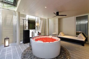 Un baño moderno con una bañera grande llena de guijarros rojos, una zona de estar con cojines y un ventilador de techo.