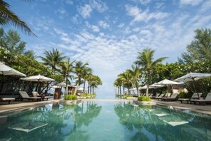 Zona de piscina con sillones de descanso y sombrillas, rodeada de palmeras, bajo un cielo despejado.
