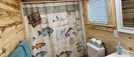 A small, wooden cabin bathroom with a shower curtain featuring fishing illustrations, a toilet, a sink, and a mirror.