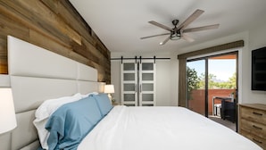 A bedroom with a bed, a ceiling fan, a sliding door to a balcony, and a TV mounted on the wall.
