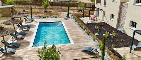 A rectangular swimming pool with lounge chairs and umbrellas, surrounded by a wooden deck and a garden area with plants and a small tree.