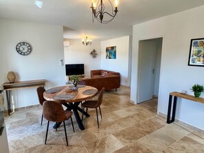 Un coin repas avec une table ronde, des chaises, un lustre, une horloge et un téléviseur en arrière-plan.