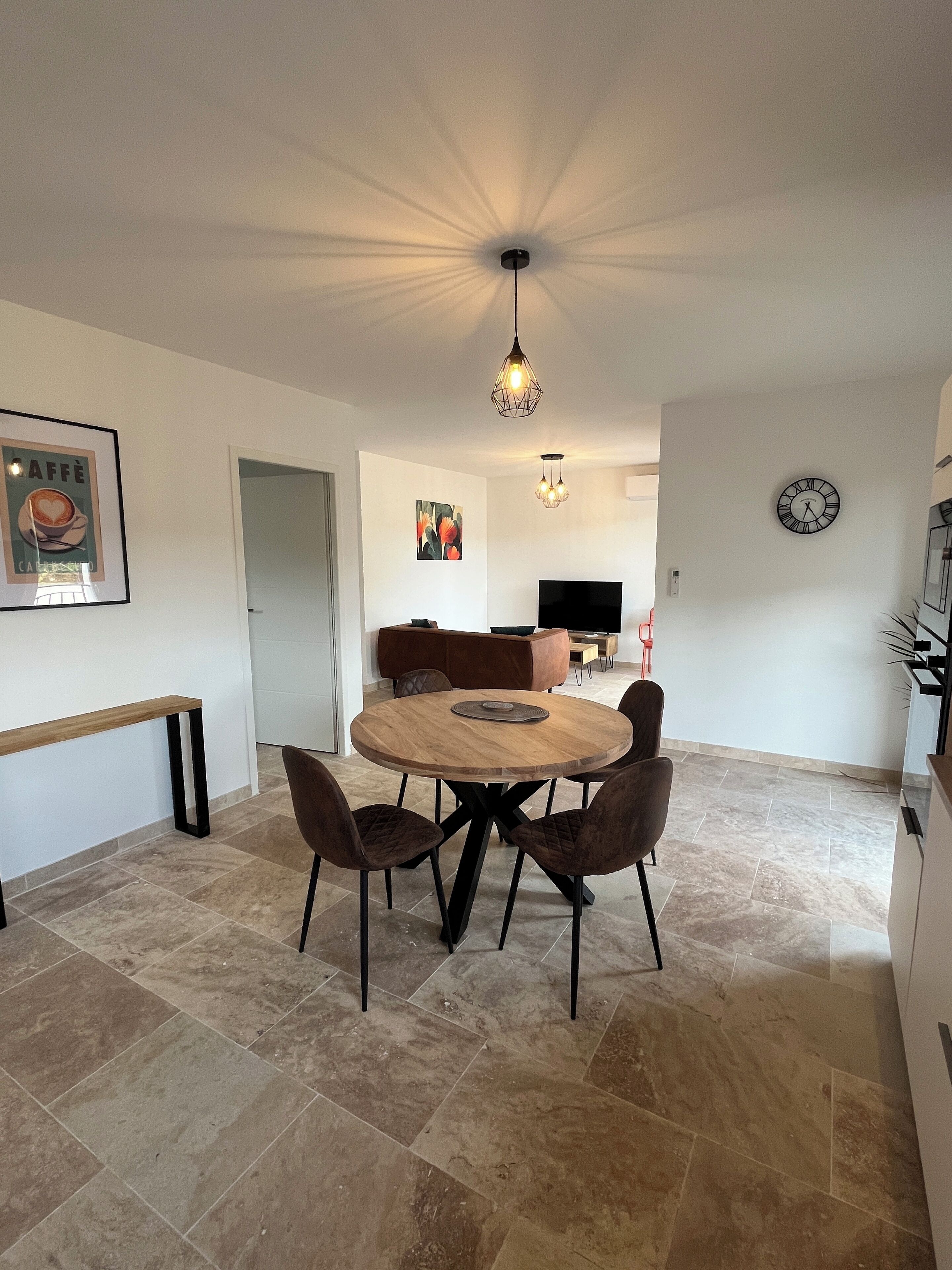 Un coin repas comprenant une table ronde en bois, quatre chaises, un banc, une horloge murale et un luminaire suspendu au plafond.