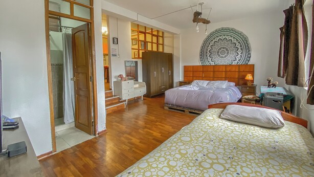 A bedroom with a wooden floor, a bed with patterned bedding, a wardrobe, a nightstand with a lamp, and a decorative wall hanging.