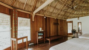Een kamer met houten wanden en een rieten dak, met een bed, een bankje en een kleine tafel met een ventilator.