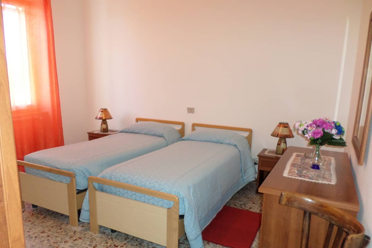 Una camera da letto con due letti singoli, un comodino con una lampada, un tavolino con un vaso di fiori e un tappeto rosso.