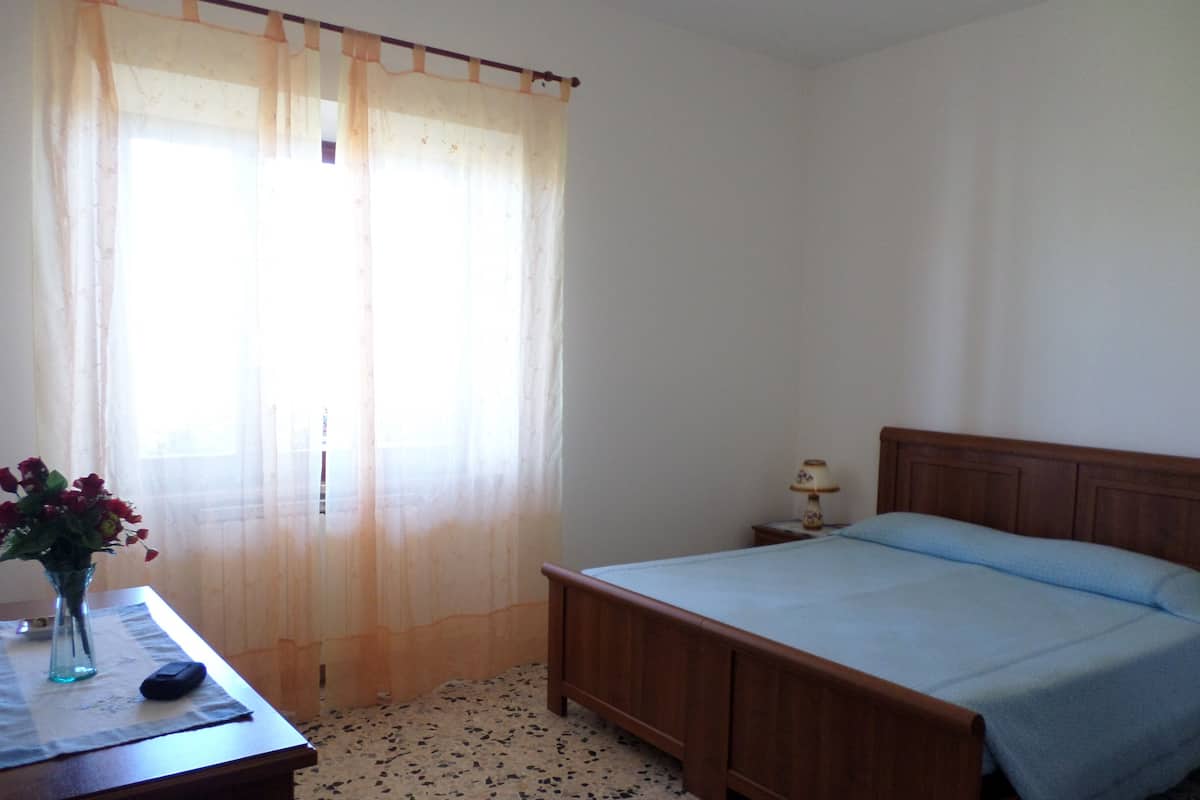 Una camera da letto con un letto di legno, un comodino con una lampada, un vaso con fiori su un tavolo e tende leggere alla finestra.