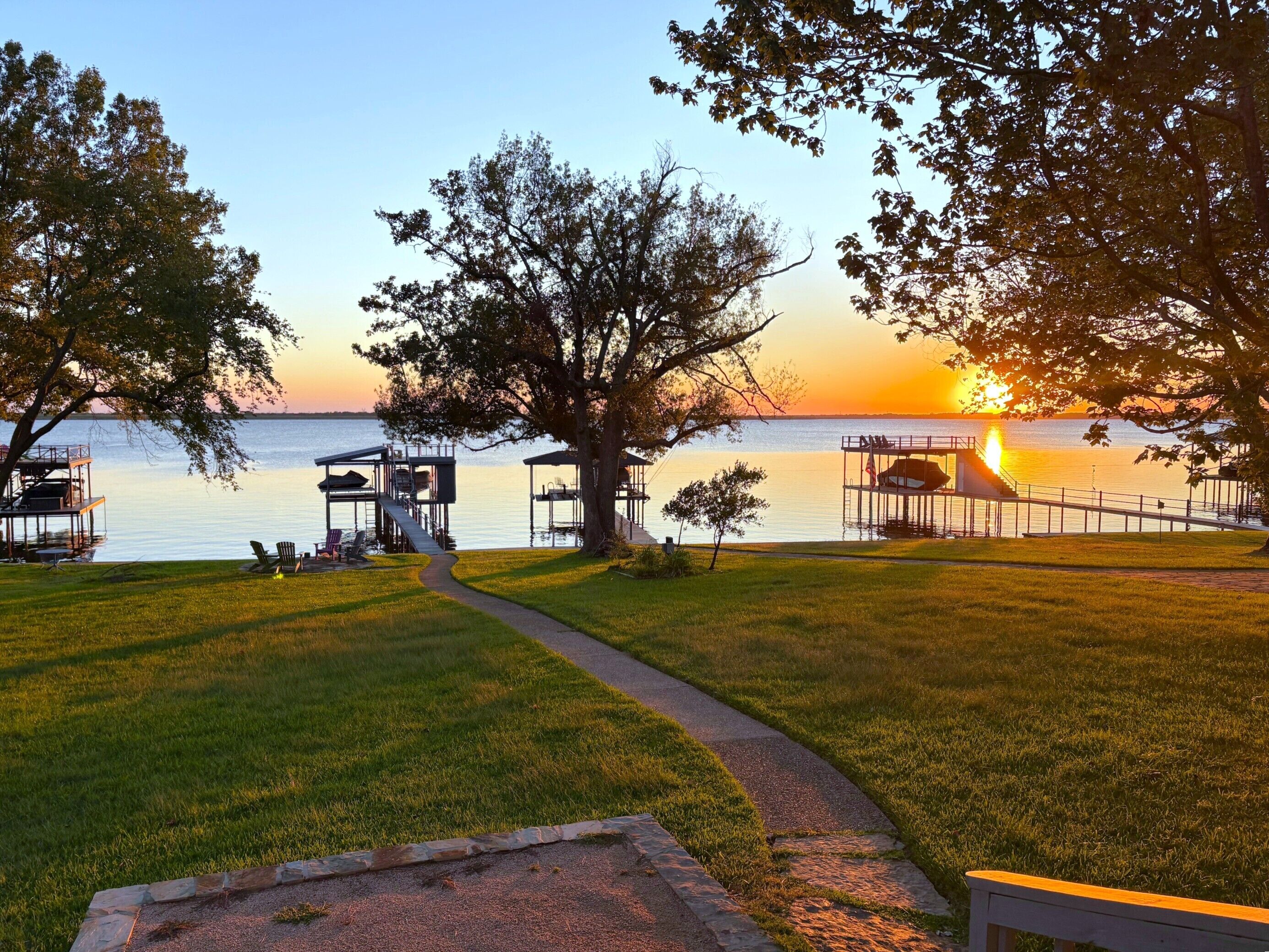 Um deck à beira do lago com uma passarela que leva a um píer, cercado por árvores e uma vista para o pôr do sol.