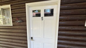 A wooden cabin with a white door marked with the number 5, a welcome sign, and a doormat with a forest scene.