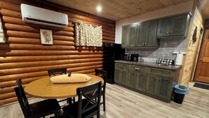 A cozy log cabin interior with a wooden dining table, black chairs, a kitchen with green cabinets, and a white air conditioner on the wall.