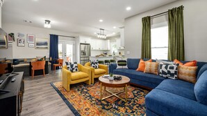 A modern living room with a blue sofa, yellow armchairs, and a coffee table. A kitchen is visible in the background with white cabinets and a refrigerator. - ATL Escape ~ Arcade I Fire pit I Patio! (Decatur)