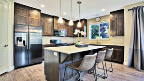 Una cocina moderna con armarios de madera oscura, electrodomésticos de acero inoxidable, iluminación colgante y un amplio ventanal con vistas a zonas verdes.