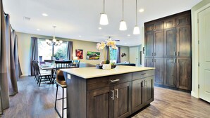 Una cocina moderna con armarios de madera oscura, una isla central con taburetes de bar, iluminación colgante y un comedor con vistas al exterior.