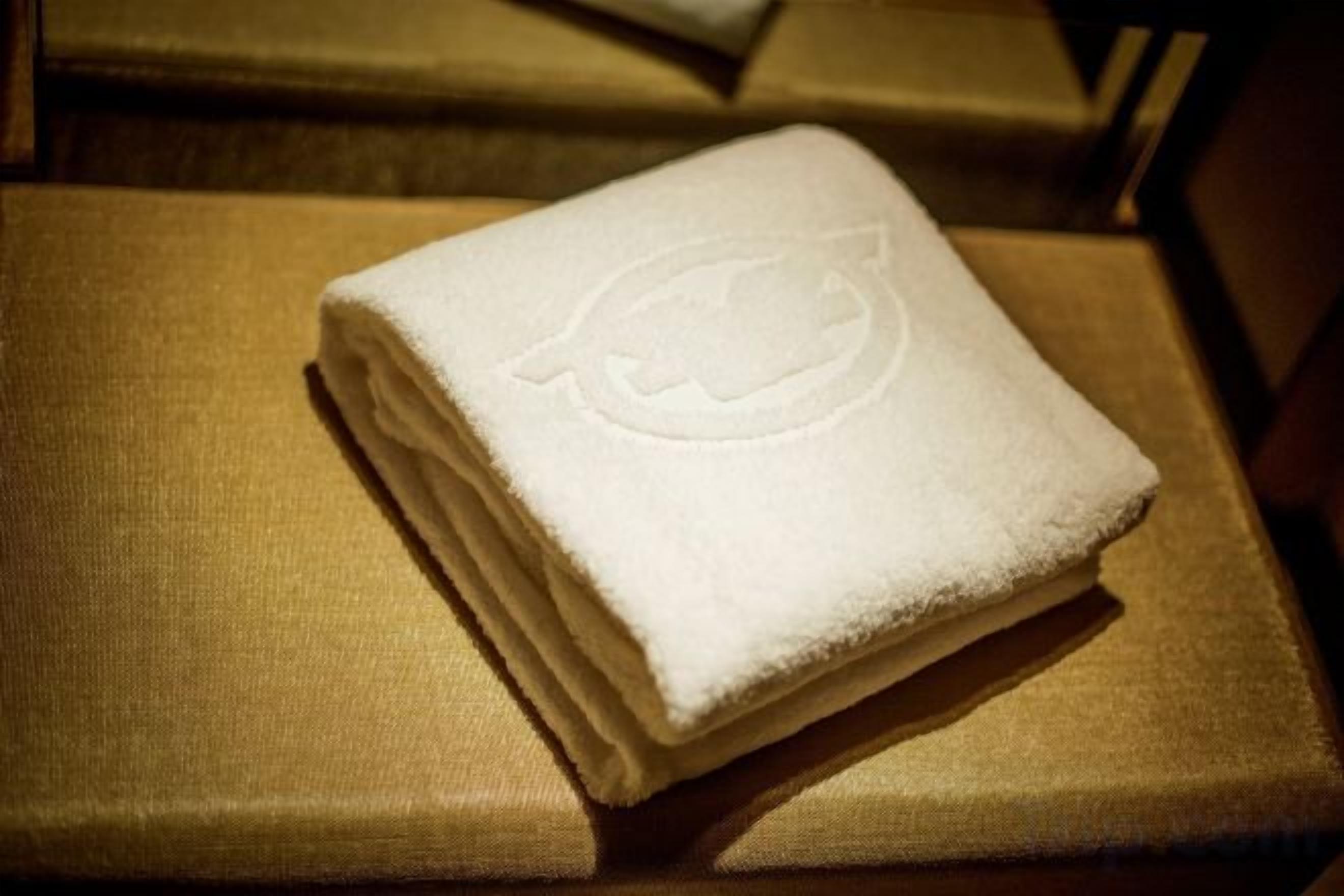 Ein gefaltetes weißes Handtuch mit einem Logo auf einer strukturierten Oberfläche, möglicherweise ein Bett oder ein Stuhl.