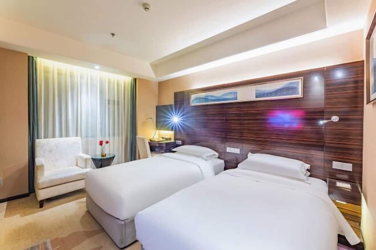 Habitación de hotel con dos camas, una silla, una mesita de noche y un ventanal con cortinas.