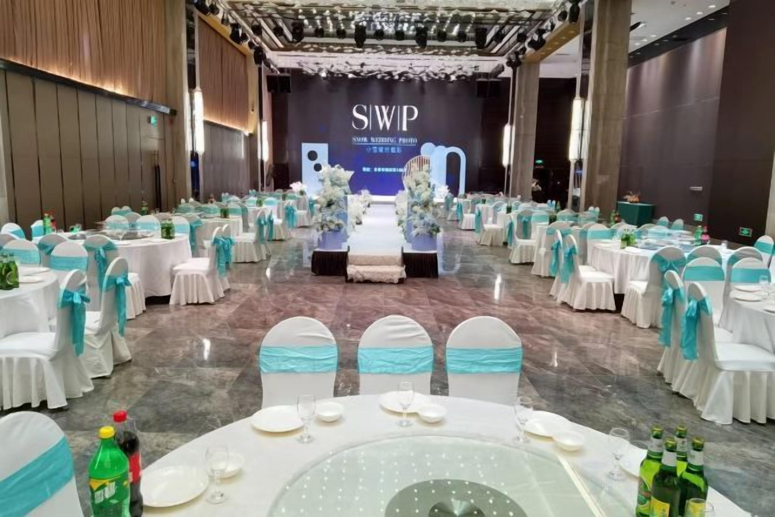A banquet hall with round tables set for a formal event, a stage with a backdrop, and a row of chairs with blue sashes.