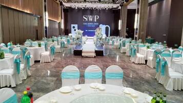 A banquet hall with round tables set for a formal event, a stage with a backdrop, and a row of chairs with blue sashes.