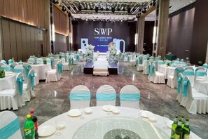 Un salón de banquetes con mesas redondas dispuestas para un evento formal, un escenario con un telón de fondo y una fila de sillas con cintas azules.
