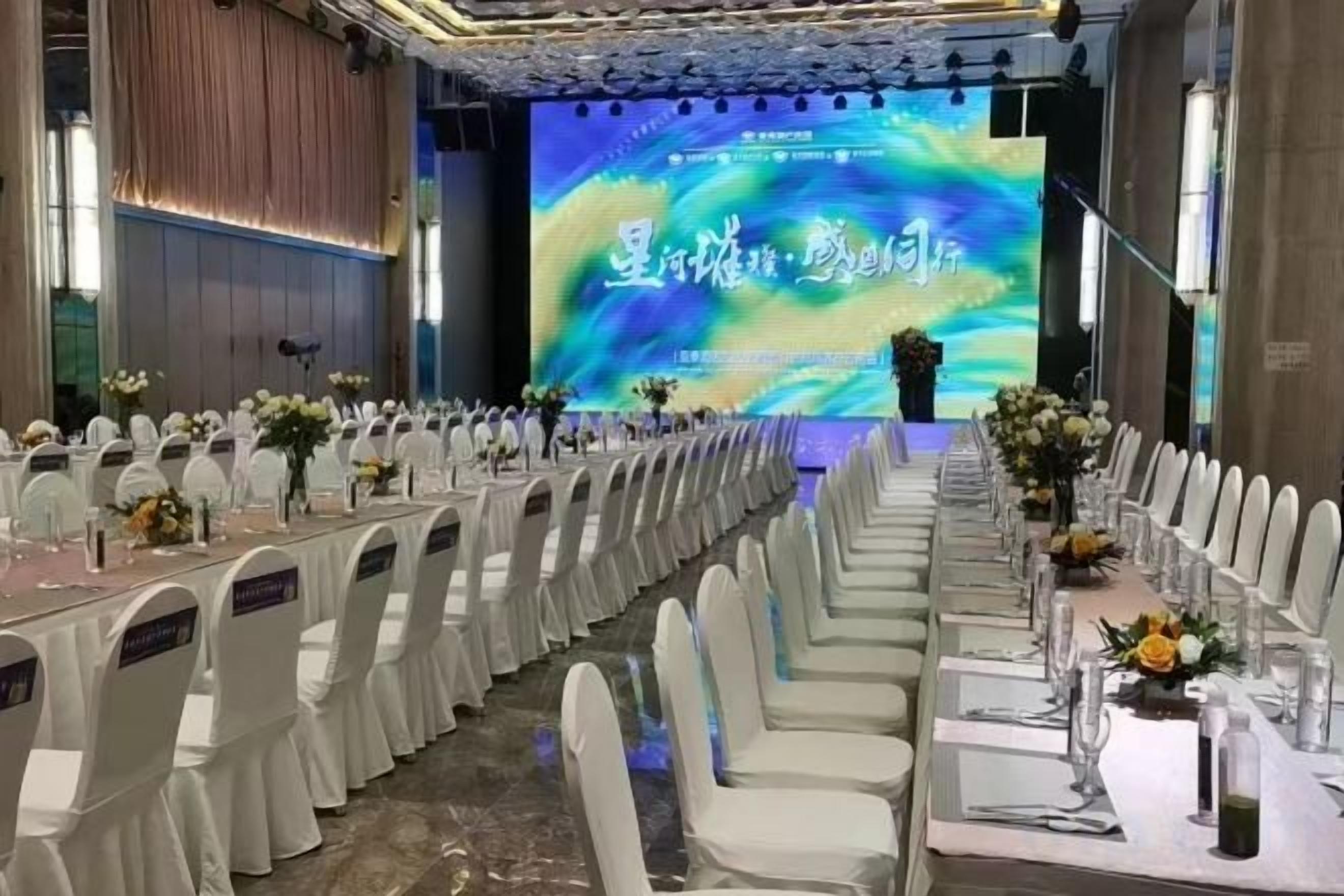 A conference room with rows of white chairs and tables, a stage with a large screen, and a formal setting.