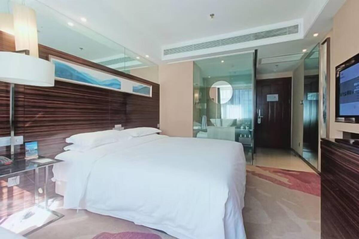 Una habitación de hotel moderna con una cama grande, un escritorio con computadora, un televisor y un muro de vidrio.