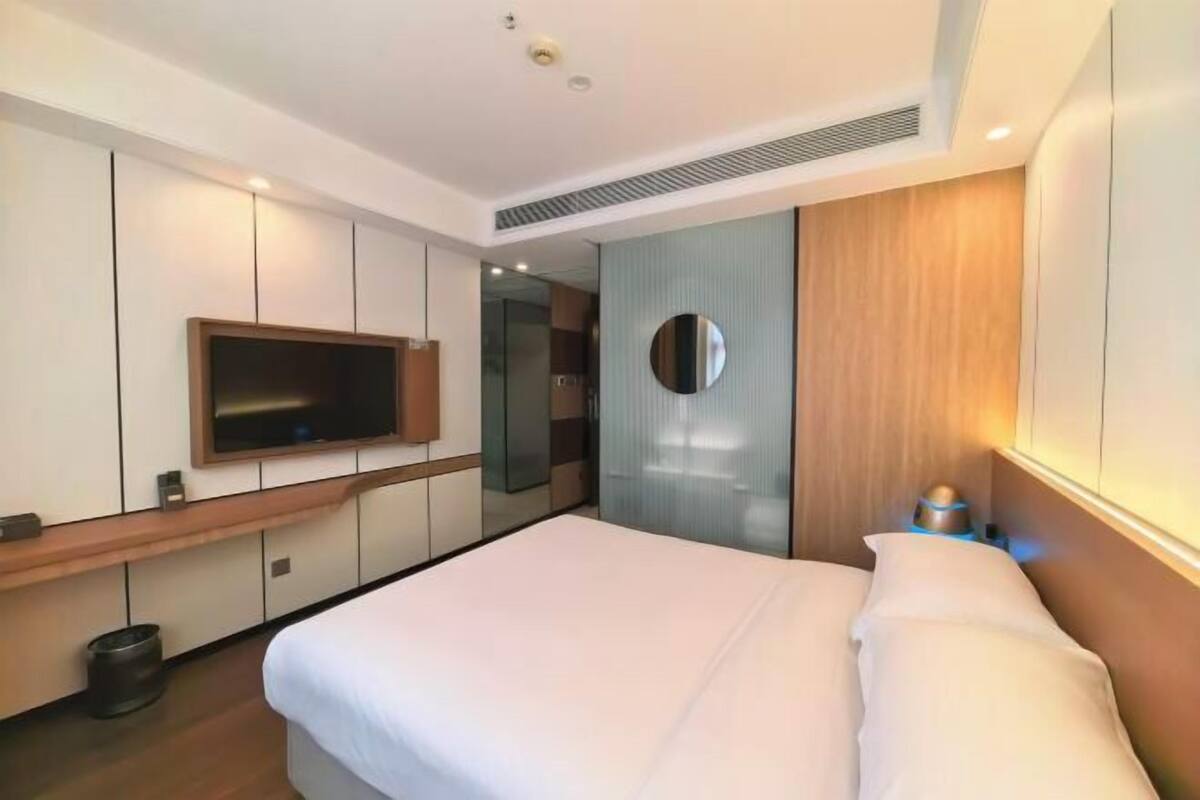 Una habitación de hotel moderna con una cama grande, un televisor de pantalla plana montado en la pared, un espejo redondo y una mesita de noche con una lámpara.