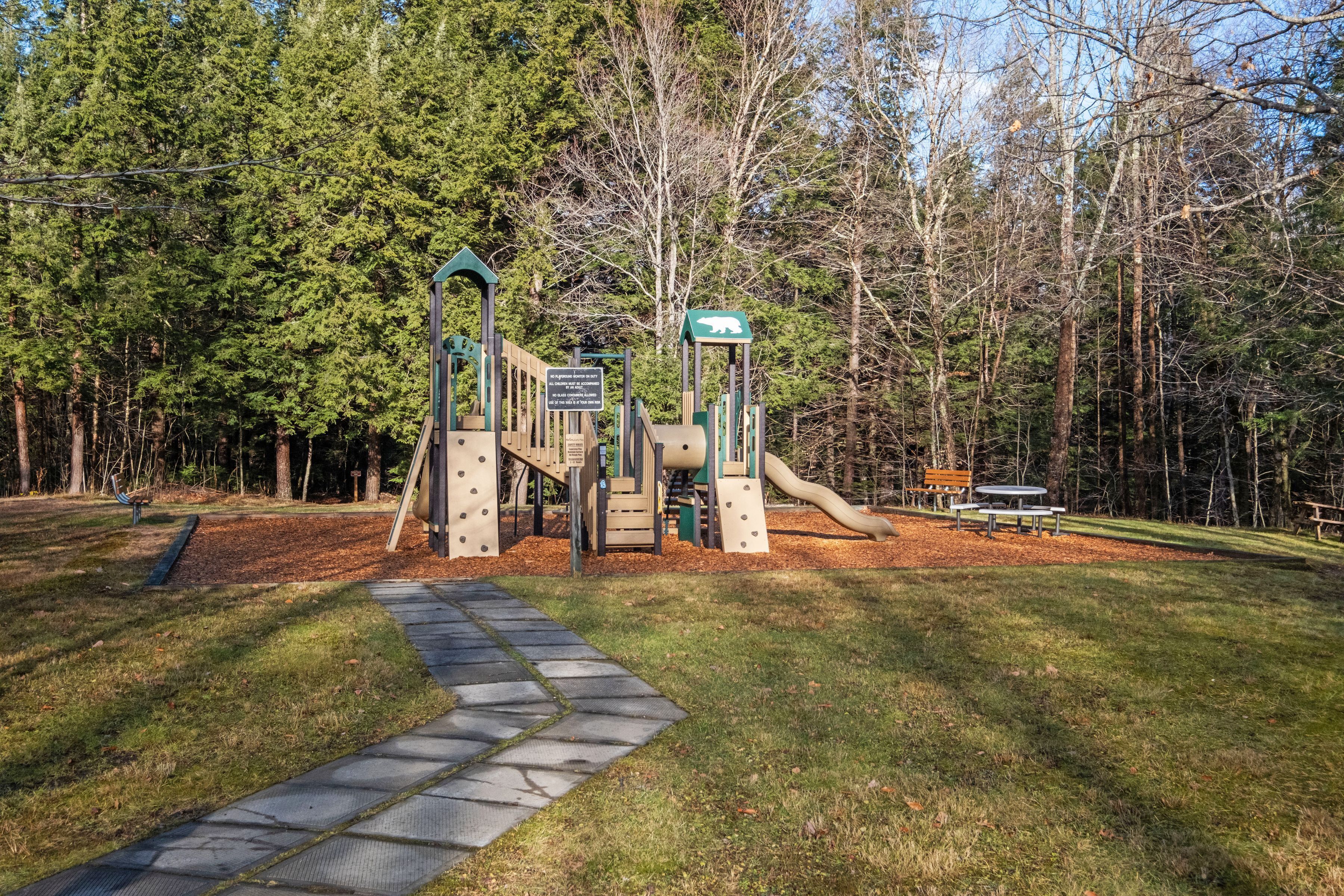Une aire de jeux avec un toboggan, des structures d’escalade et une table de pique-nique, située dans un parc arboré.
