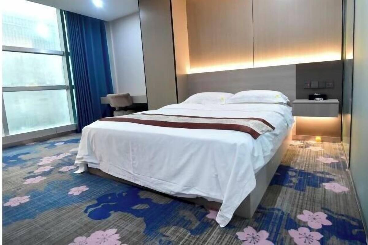 Una habitación de hotel con cama, mesita de noche, ventana con cortinas y alfombra estampada con motivos florales.