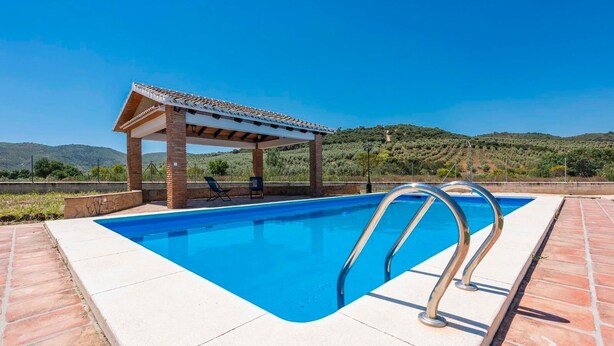Una piscina rettangolare con una pergola e lettini prendisole, circondata da un solarium piastrellato e uno sfondo montuoso.