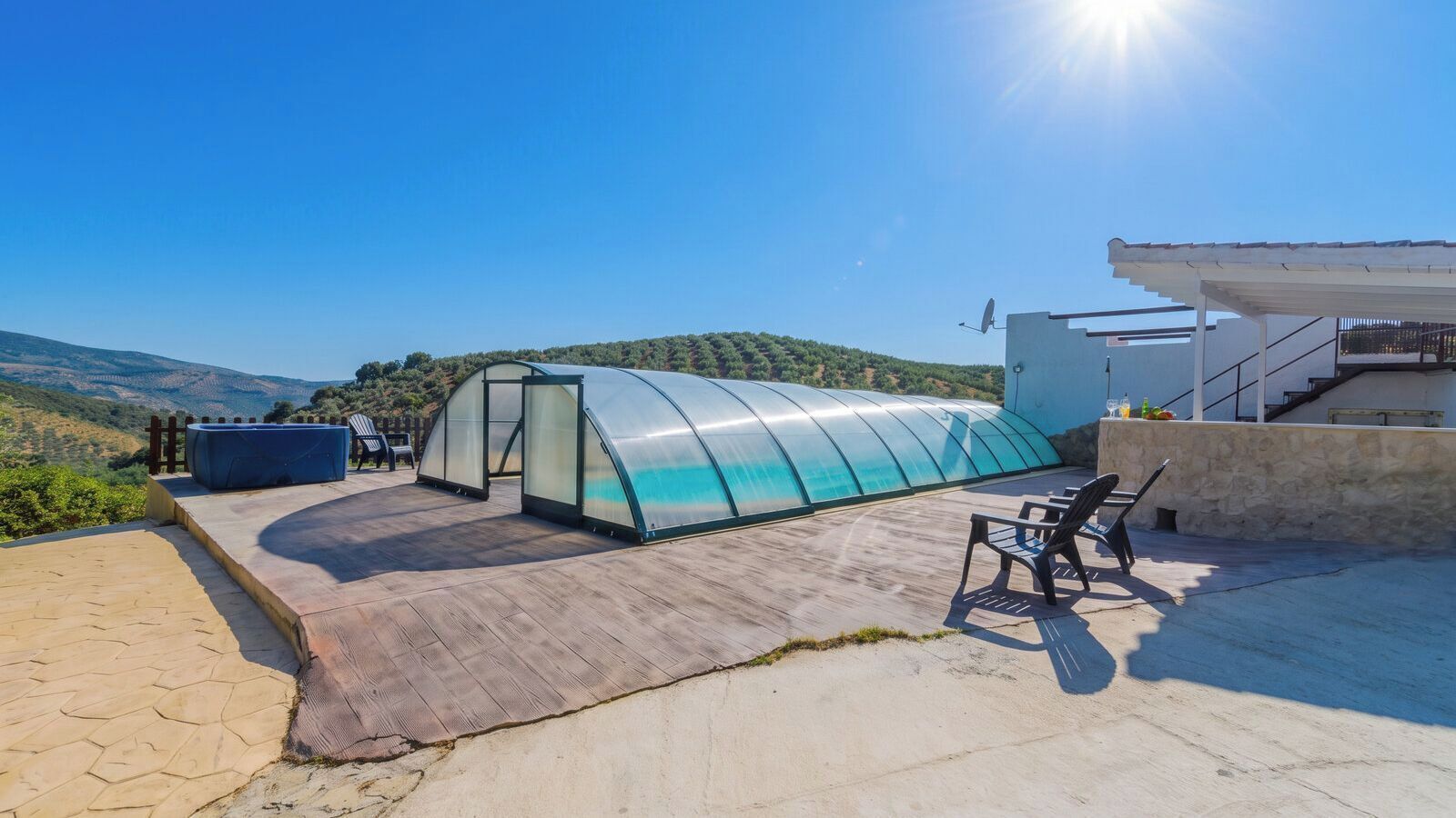 Eine Gewächshauskonstruktion mit gewölbter Dachform, eine Terrasse mit Sitzgelegenheiten und Blick auf Hügel.