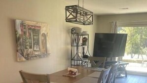 A dining area with a wooden table and chairs, a hanging light fixture, a painting on the wall, and a TV in the background.