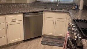 A compact kitchen with white cabinets, a built-in dishwasher, a stove, and a window with blinds.