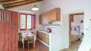 A rustic kitchen with wooden cabinets, a dining table with chairs, and a window with pink shutters. - El Alambique Frigiliana by Ruralidays (Málaga)
