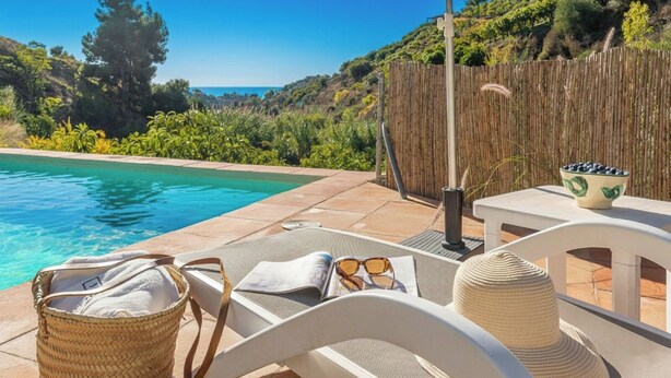 A poolside area with a wicker basket, a white chair, a table with a bowl of fruit, and a view of the sea. - El Alambique Frigiliana by Ruralidays (Málaga)