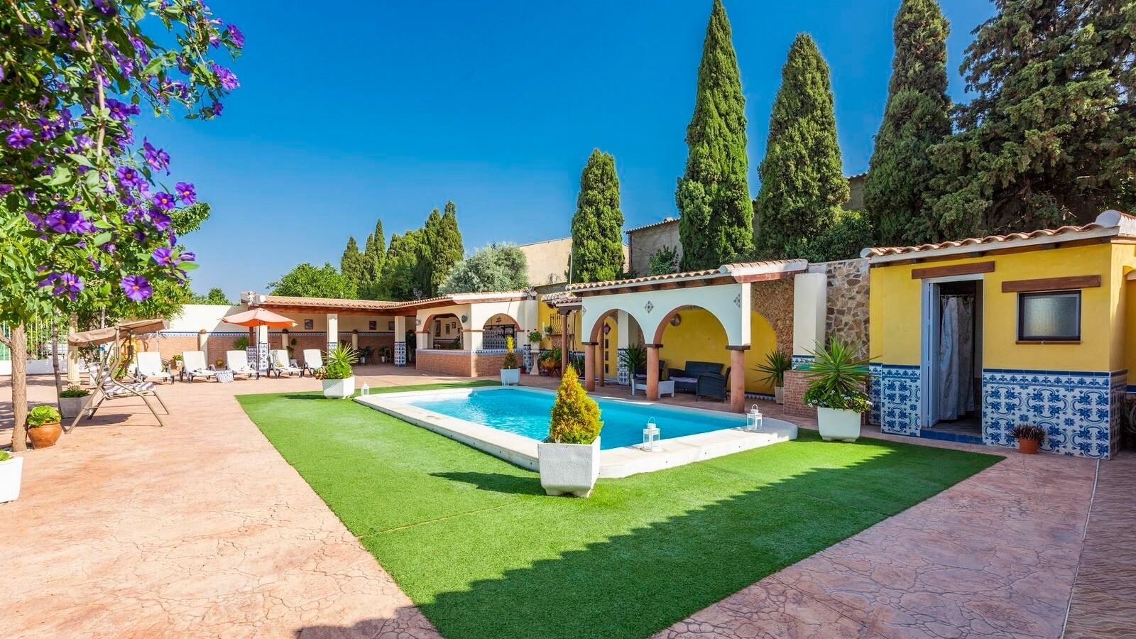 Un espace piscine doté d’une piscine rectangulaire, entouré d’une terrasse avec des chaises et une petite table. On aperçoit en arrière-plan de grands arbres et un bâtiment avec un toit recouvert de tuiles.