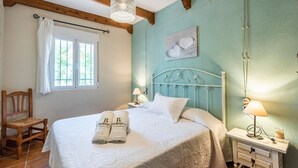 A neatly made bed with white linens and two towels with a logo, a wooden chair, a bedside table with a lamp, and a window with white curtains. - La Estaquilla Dúrcal by Ruralidays (Granada)