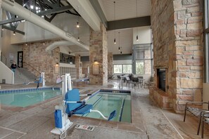 An indoor swimming pool with a stone wall and a fireplace, surrounded by seating and a staircase.