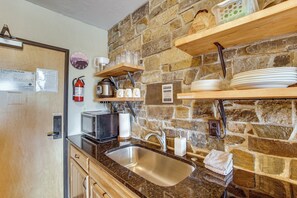 A kitchen with a stone wall, a sink, a microwave, a fire extinguisher, and a fire escape plan on the door.