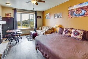 A room with a bed, a sofa, a dining table with chairs, a window with curtains, and artwork on the walls.
