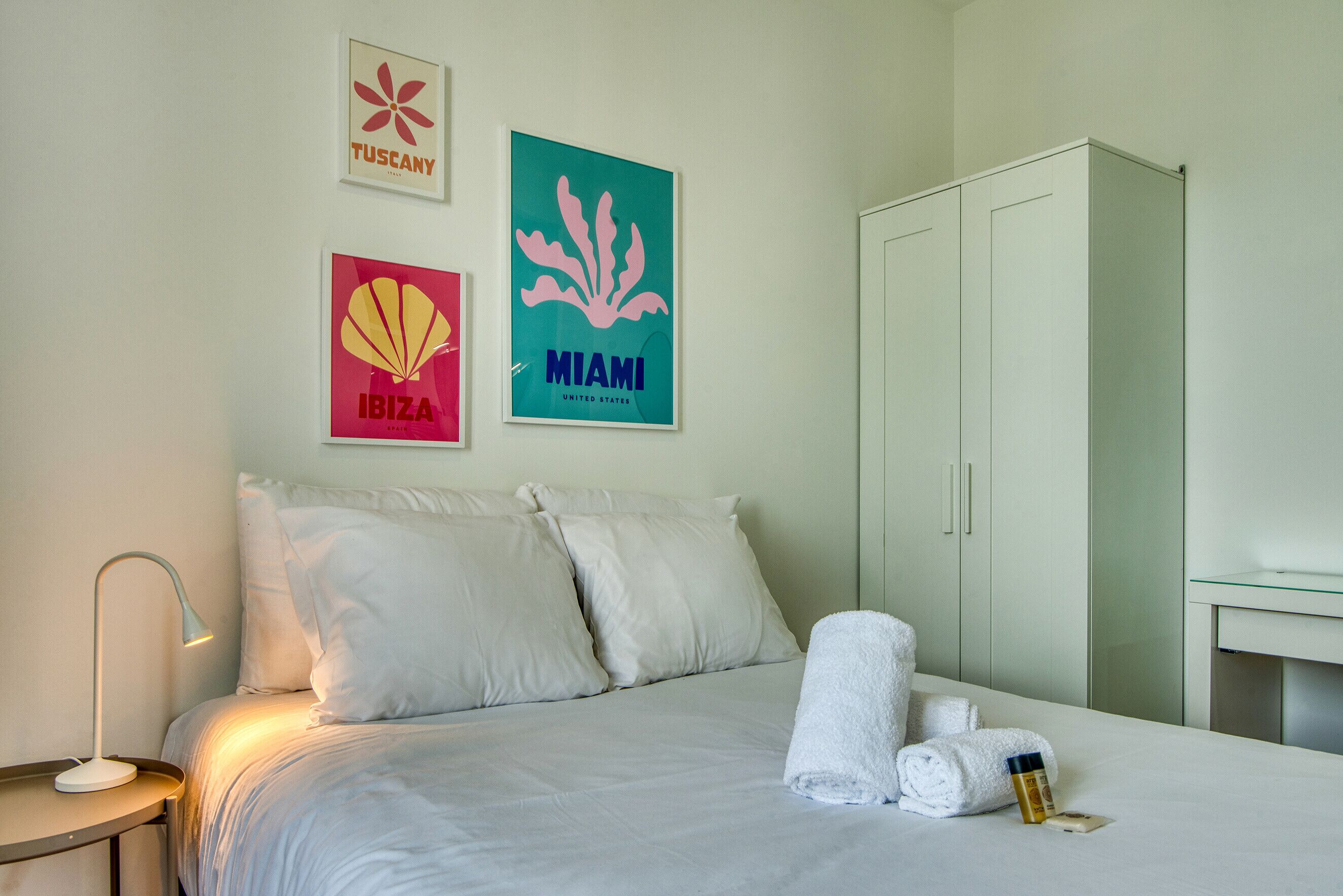 Ett hotellrum med en säng, kuddar, ett sängbord med en lampa, en garderob och tavlor på väggen som föreställer Toscana, Ibiza och Miami.