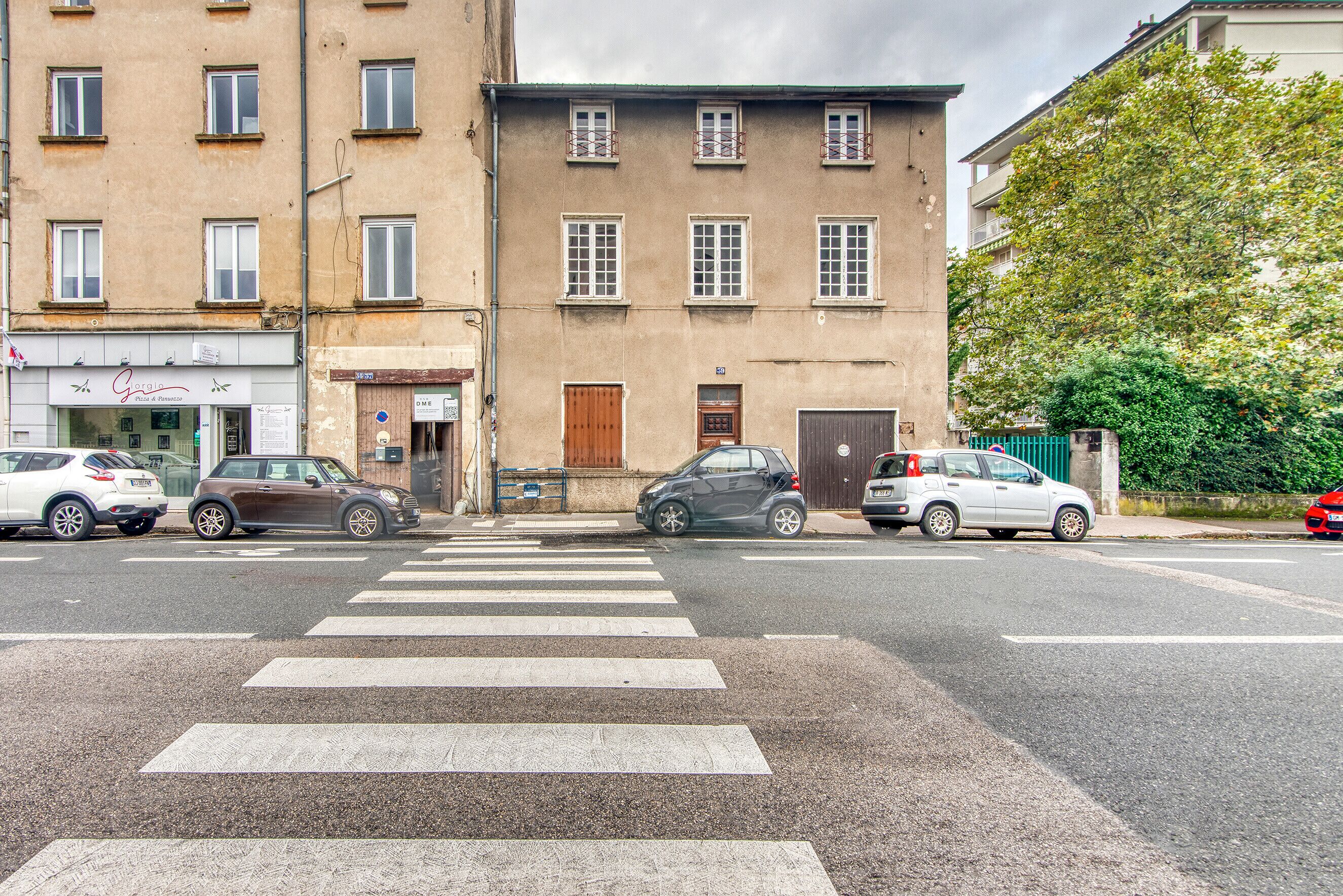 Vista de una calle con un paso de cebra, coches estacionados y edificios con escaparates.