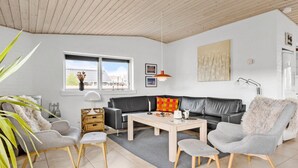 A modern living room with a black leather sofa, grey armchairs, a wooden coffee table, and a large window with a view of buildings. - Lovely home in Blokhus with WiFi (Blokhus)