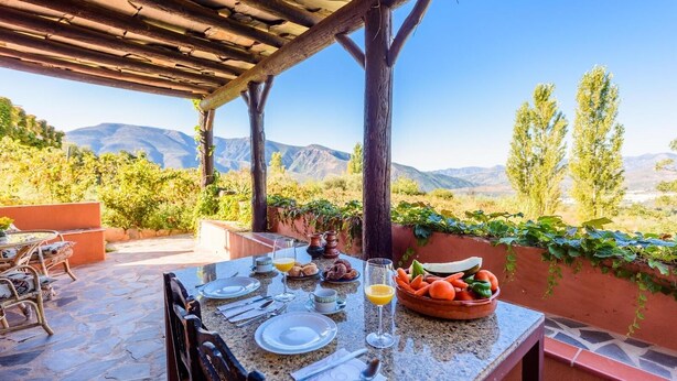 An outdoor dining area with a table set for breakfast, surrounded by mountains and greenery. - Cortijo El Cerrillo Órgiva by Ruralidays (Granada)