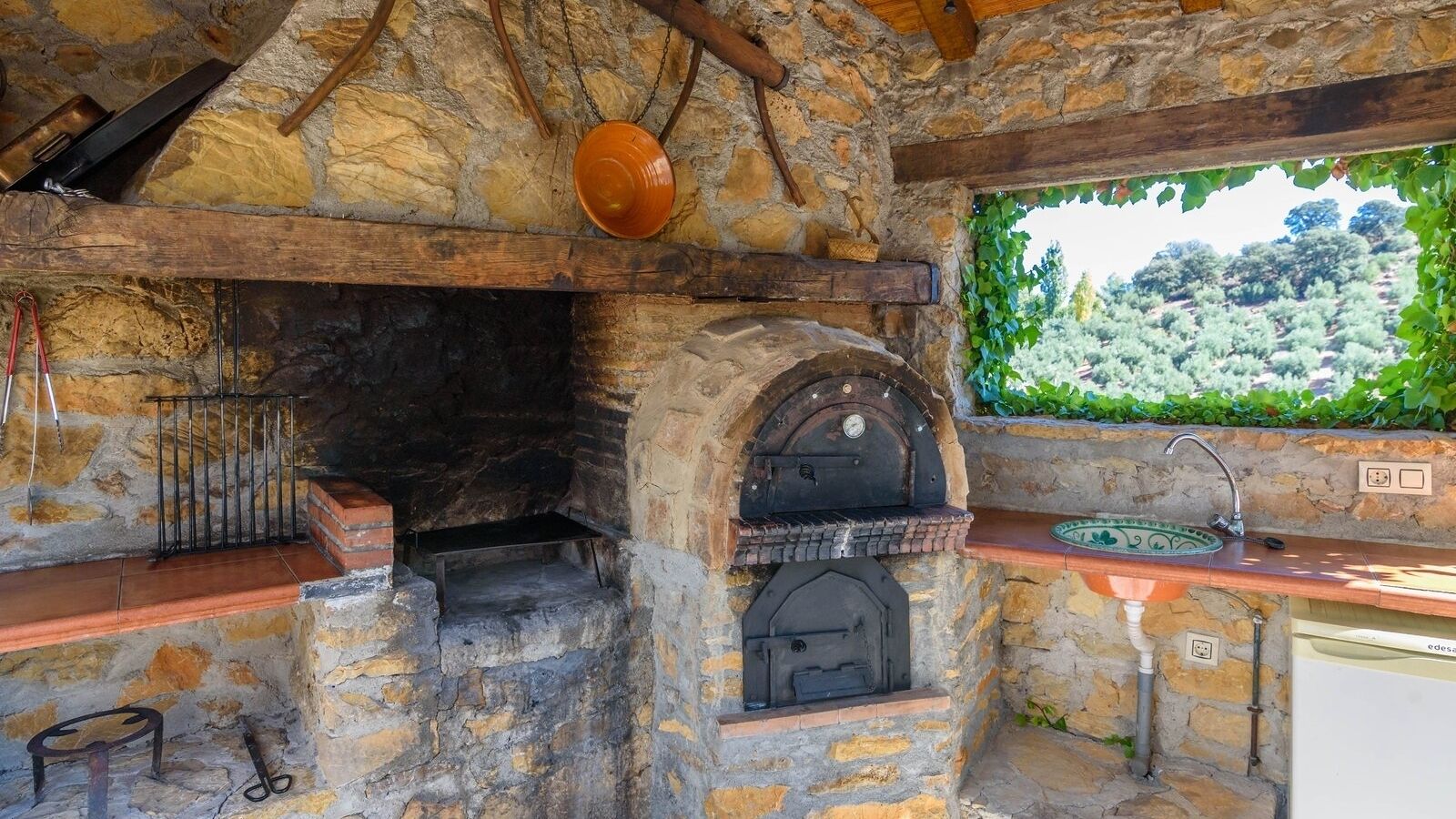Cozinha de pedra com forno a lenha, suporte para panelas pendurado e vista para um vinhedo.