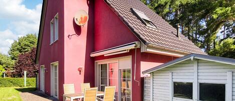 Une maison rouge à deux étages avec un petit garage attenant, une terrasse avec des meubles d’extérieur et une antenne parabolique.