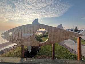 A wooden fish sculpture with a circular window, set on a grassy area with a view of a distant landscape.