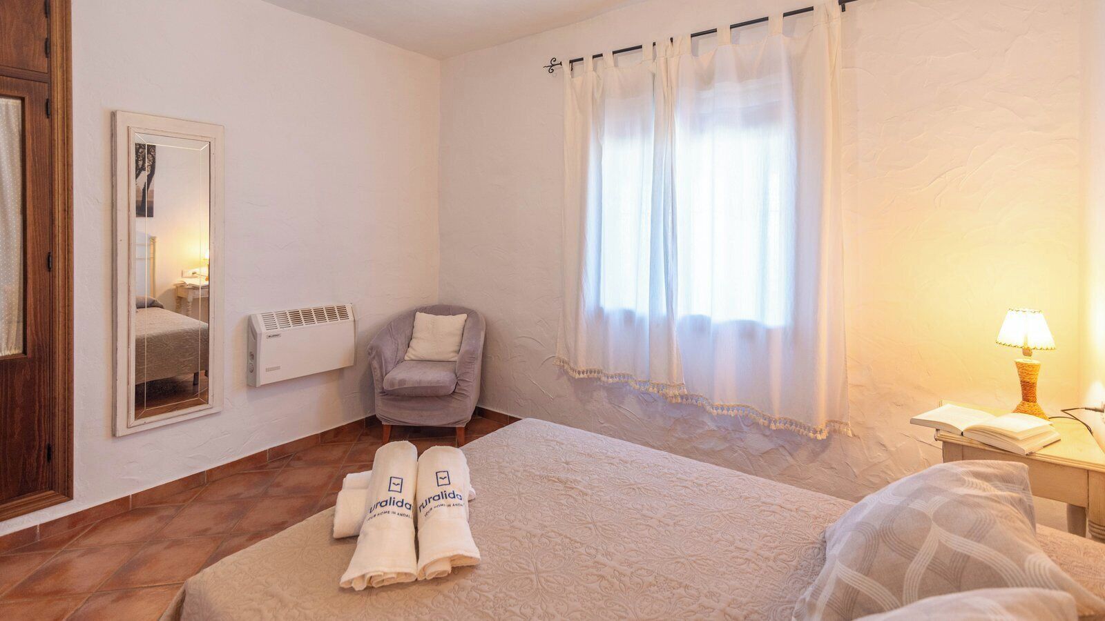 A hotel room with a bed, a chair, a window with curtains, a bedside lamp, and a mirror.