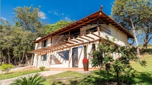 A small, single-story building with a tiled roof and wooden beams. It has a balcony with a railing and is surrounded by greenery. - Vila do Frade (Alexânia)