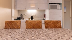 Angolo cottura compatto con pensili bianchi, forno a microonde e lavello. Due sedie con cuscini a fantasia e un letto con copripiumino dal motivo geometrico.