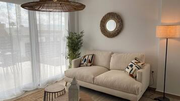 Een woonkamer met een beige bank, een ronde spiegel, een hanglamp van riet, een vloerlamp en een kleine tafel met een lantaarn.
